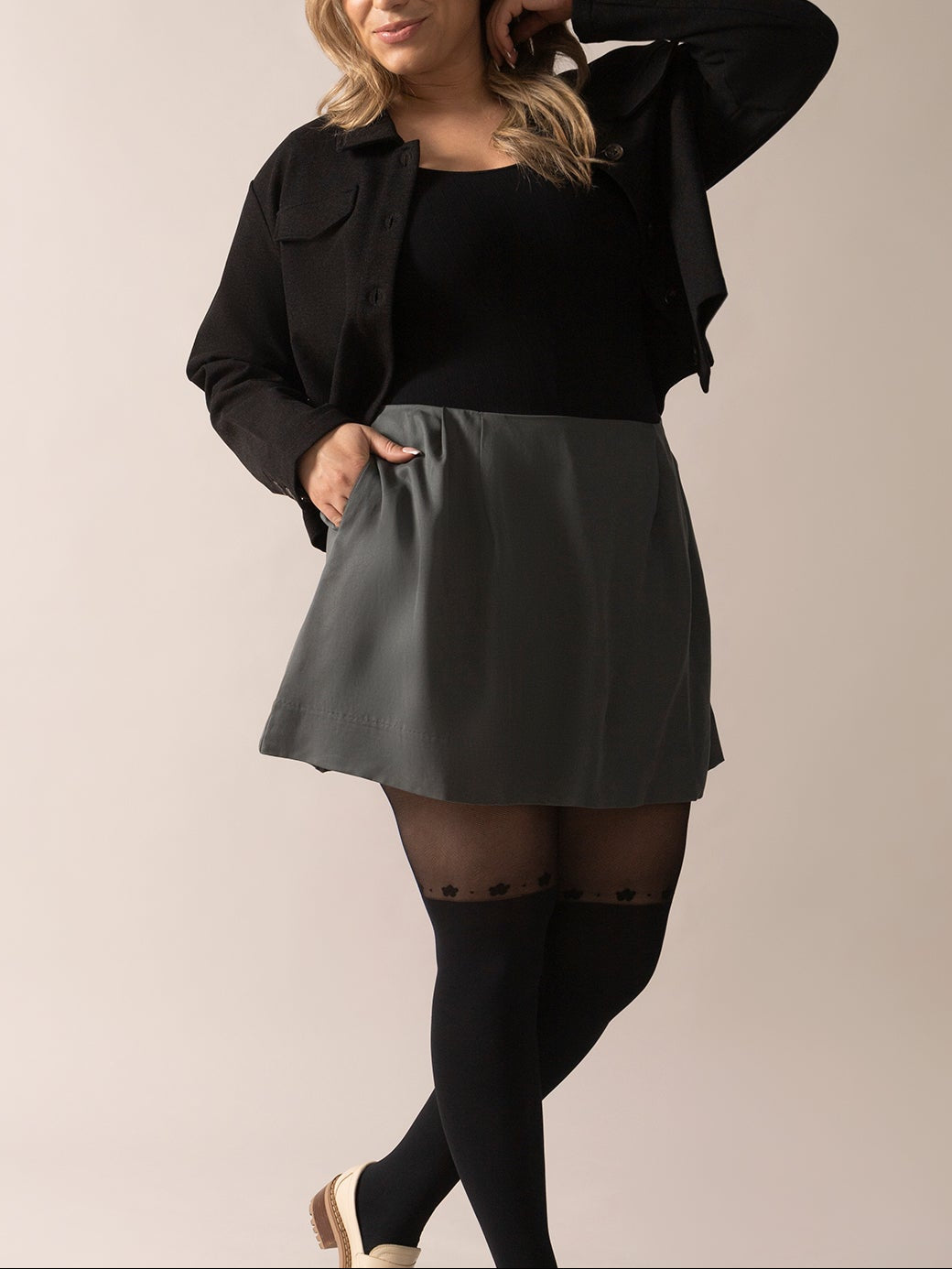 A woman posing in an A-line eco twill OTK skirt in stormy green color, paired with a black top and tights, standing against a neutral background.